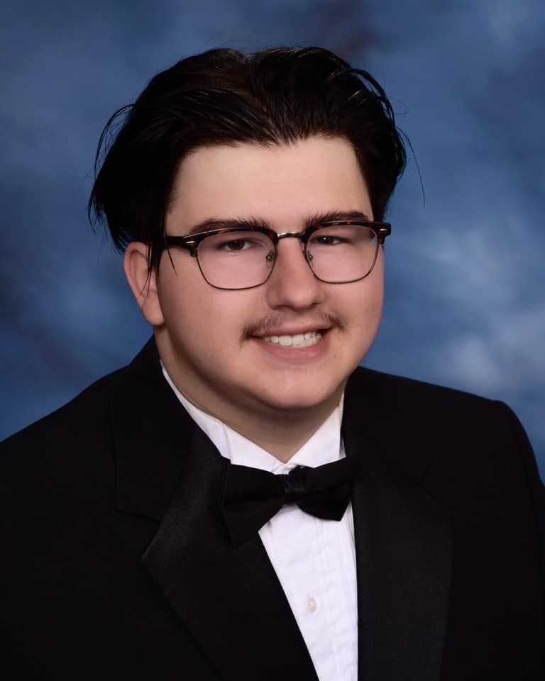 Young man in black tuxedo and bow tie with glasses smiling for formal portrait photo.