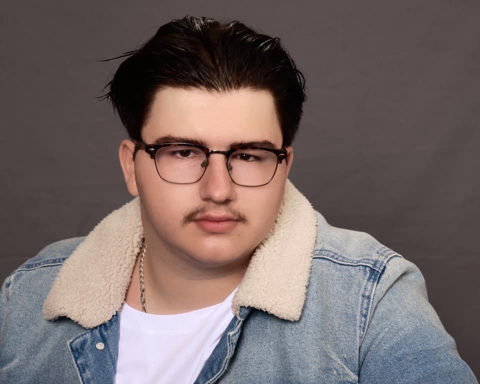 Young man with glasses and mustache wearing a denim sherpa jacket poses for a professional headshot photo.