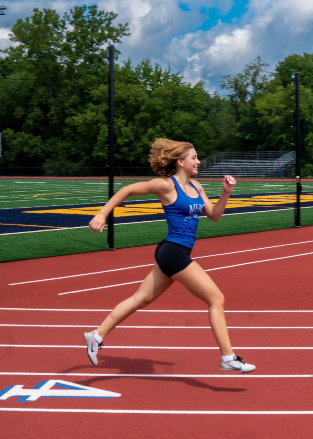 A young female track athlete in a blue jersey sprints on a red running track during an outdoor meet.