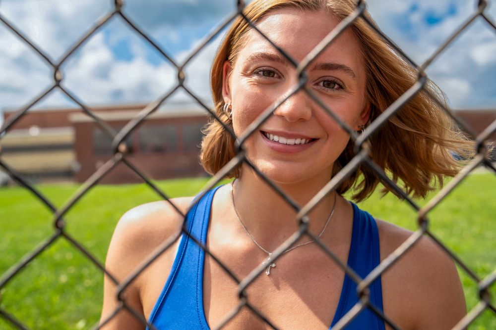 Smiling young woman in blue tank top posing behind a chain-link fence outdoors on a sunny day.