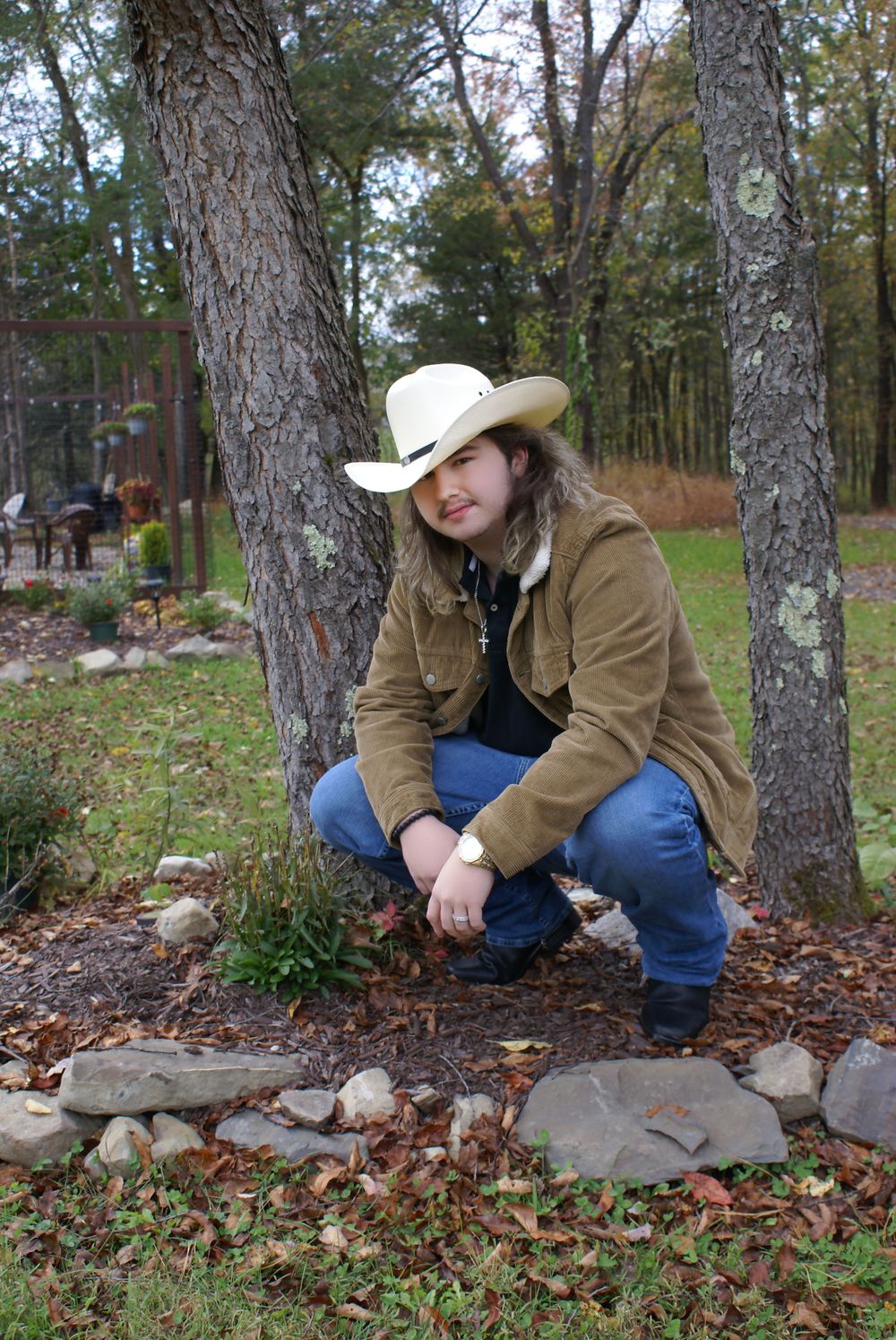 Young woman in white cowboy hat and brown jacket crouching near rocks and trees in a rural outdoor setting.