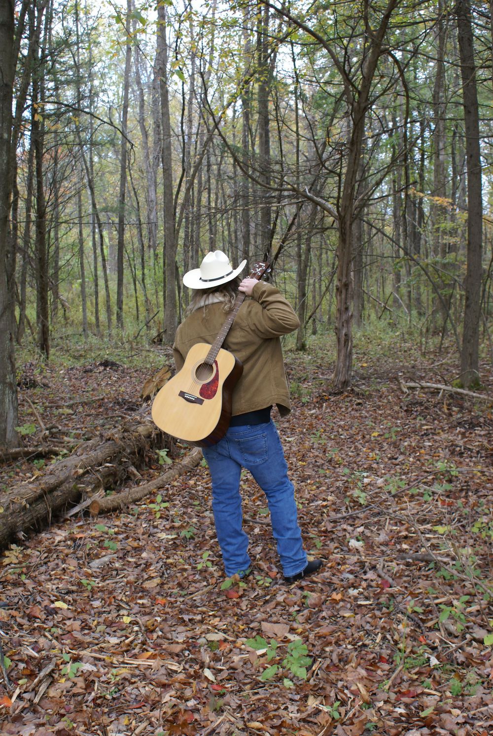 A country musician in a white cowboy hat and jeans carries an acoustic guitar through an autumn forest.