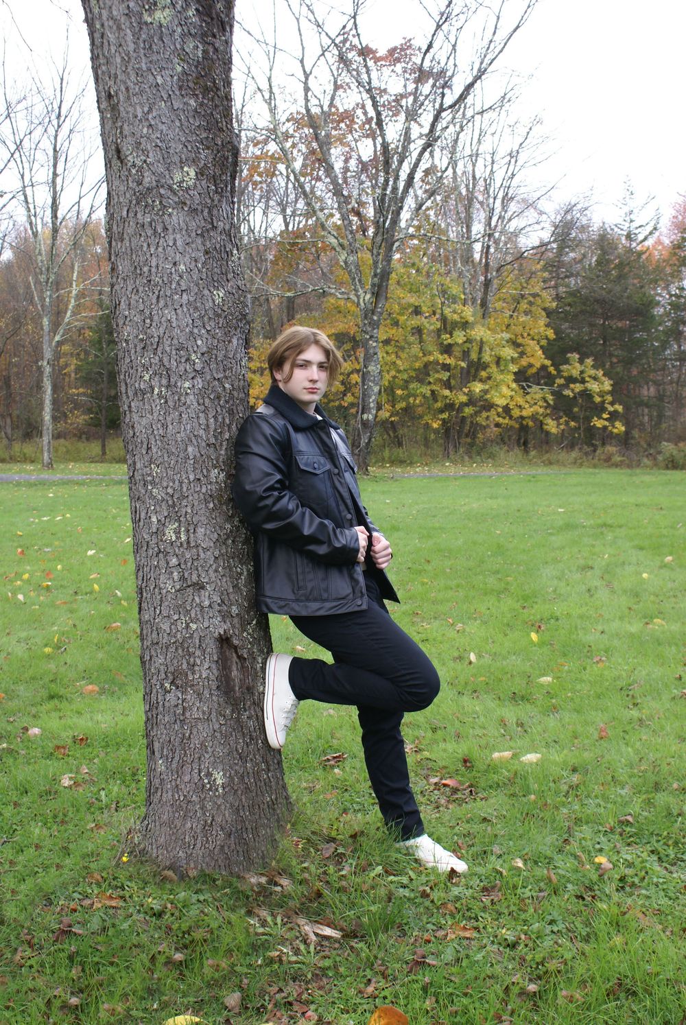 Young man in black leather jacket and jeans leaning against a tree in an autumn park with colorful foliage.