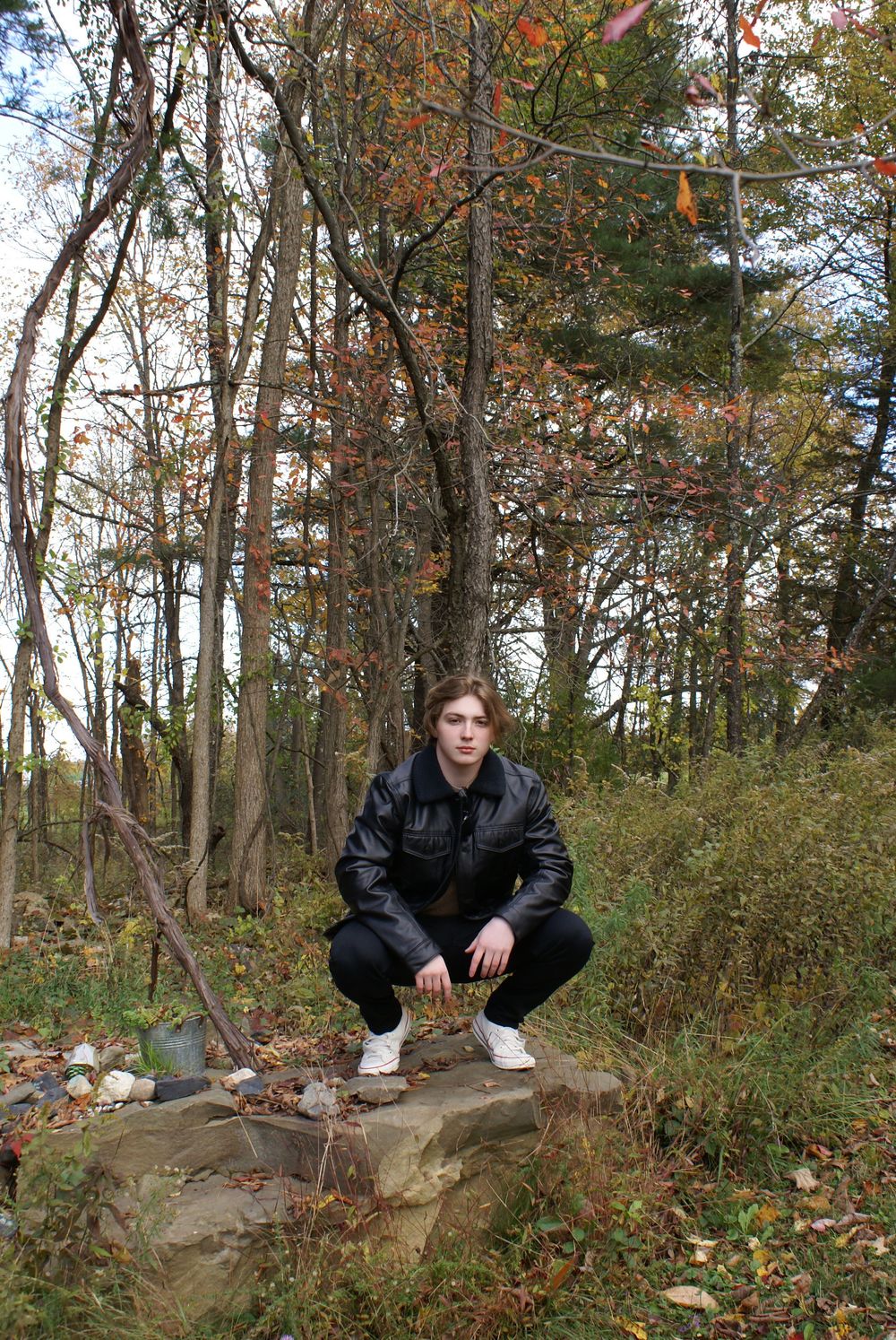 Young man in black leather jacket crouching on a rock in an autumn forest setting.