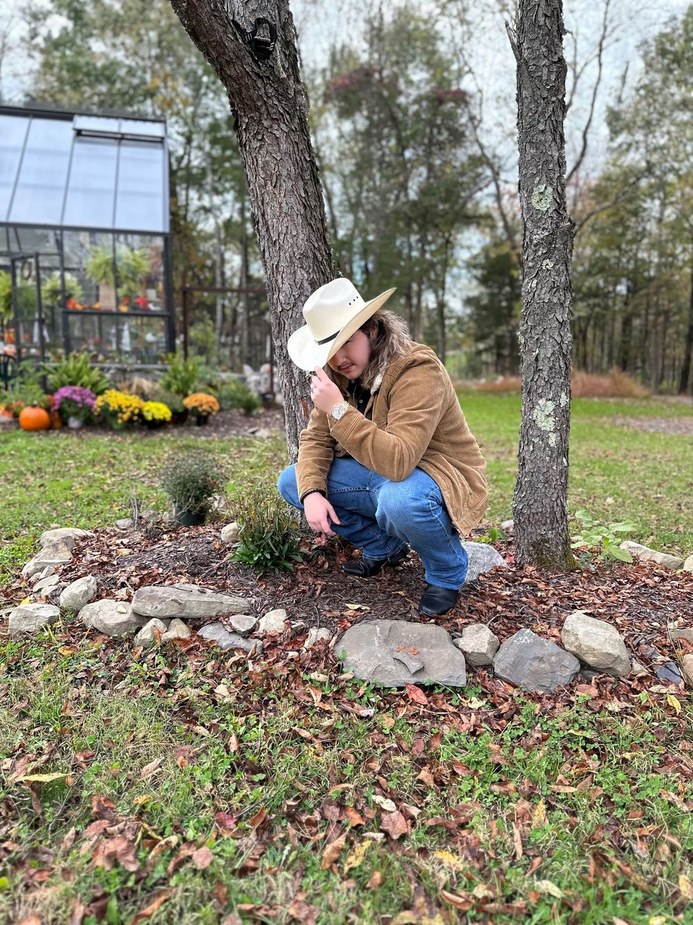 Man in cowboy hat crouching by a rock-bordered garden bed under trees, with a greenhouse visible in the background.