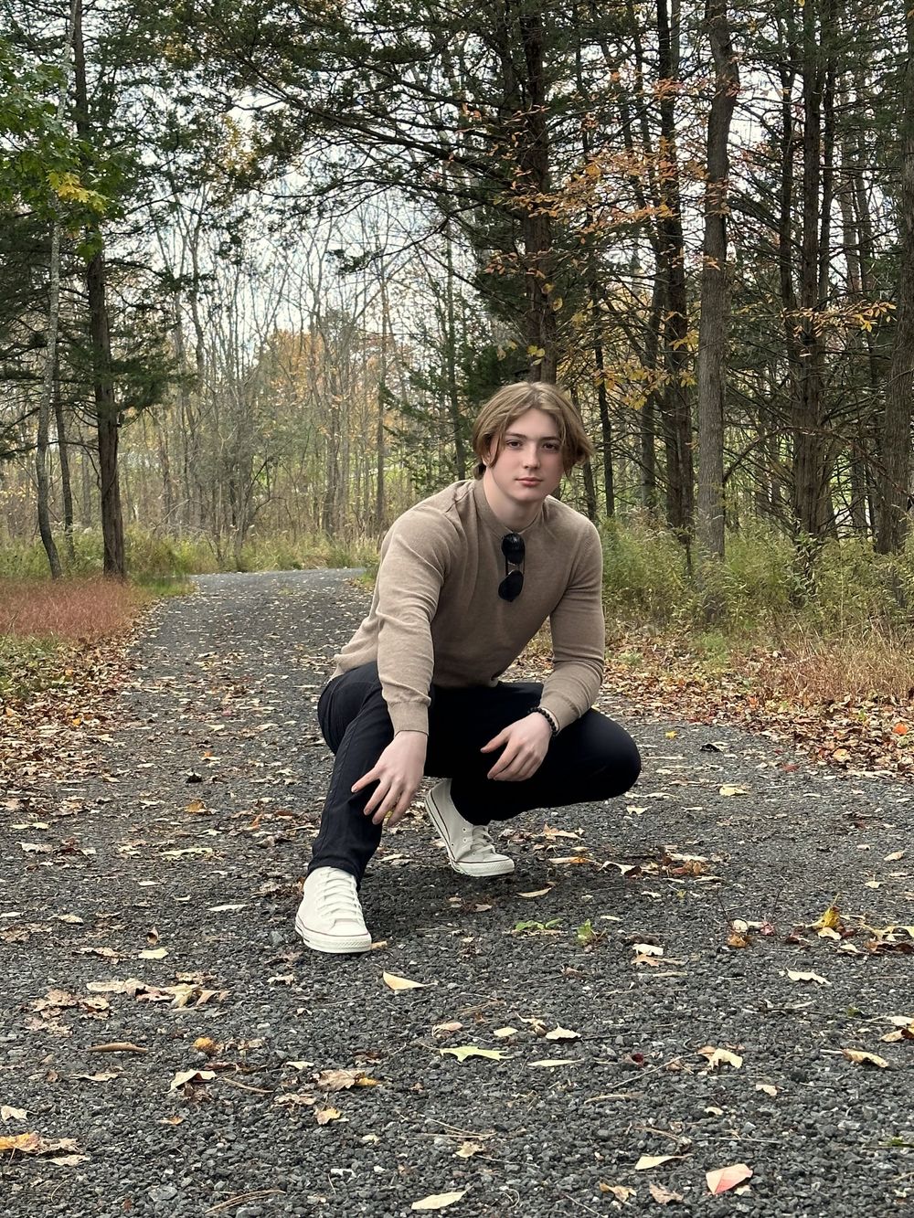 Young man crouching on a gravel path surrounded by autumn foliage, wearing a beige sweater, black jeans, and white sneakers.