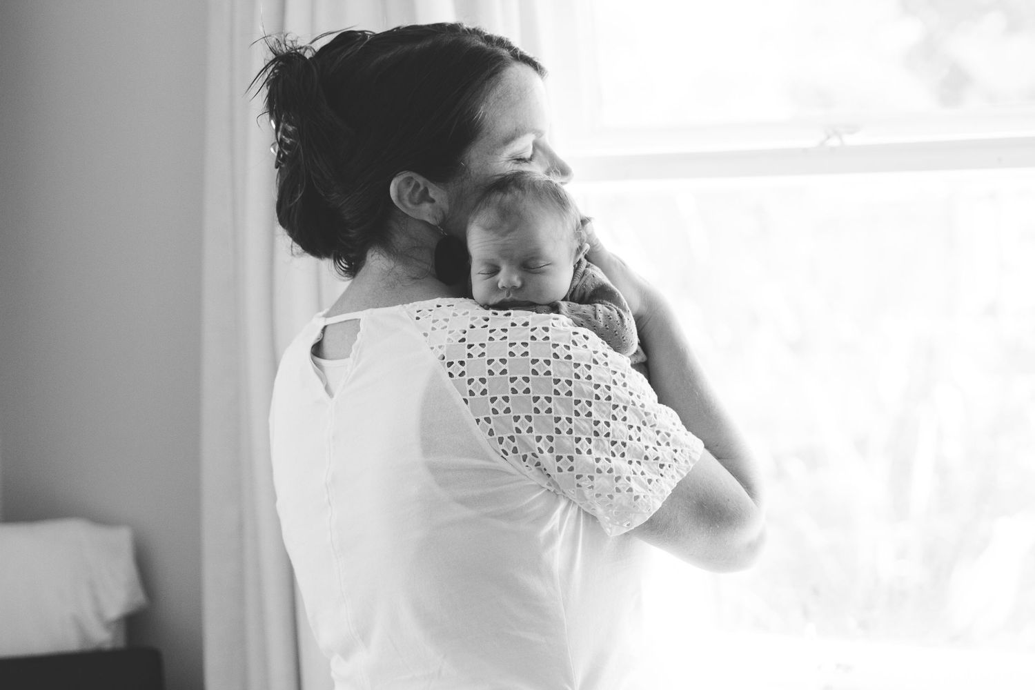 Black and white photo of a mother cradling her newborn baby on her shoulder near a bright window.