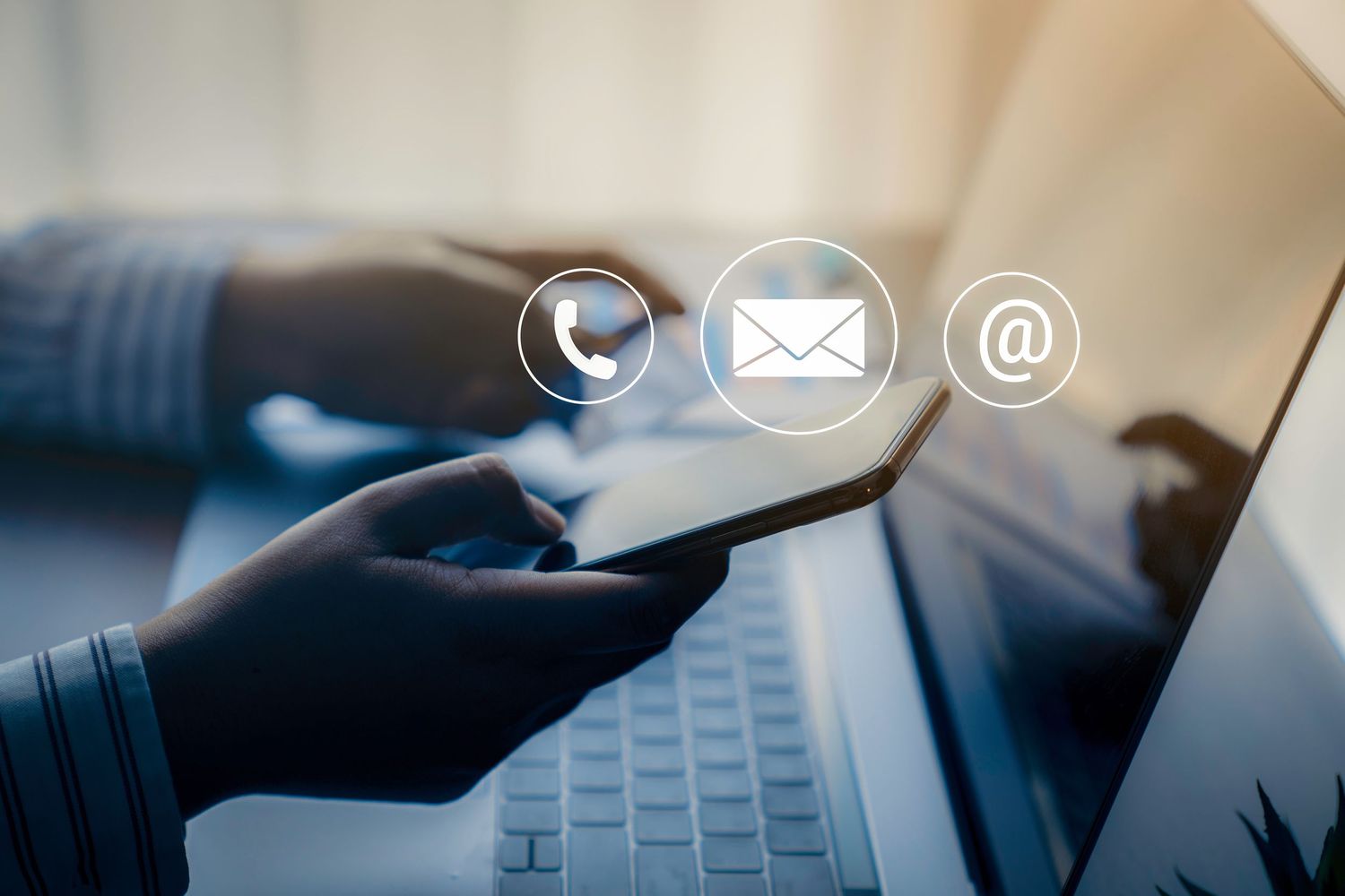 Person holding smartphone near laptop with phone, email, and at-sign contact icons floating above device.