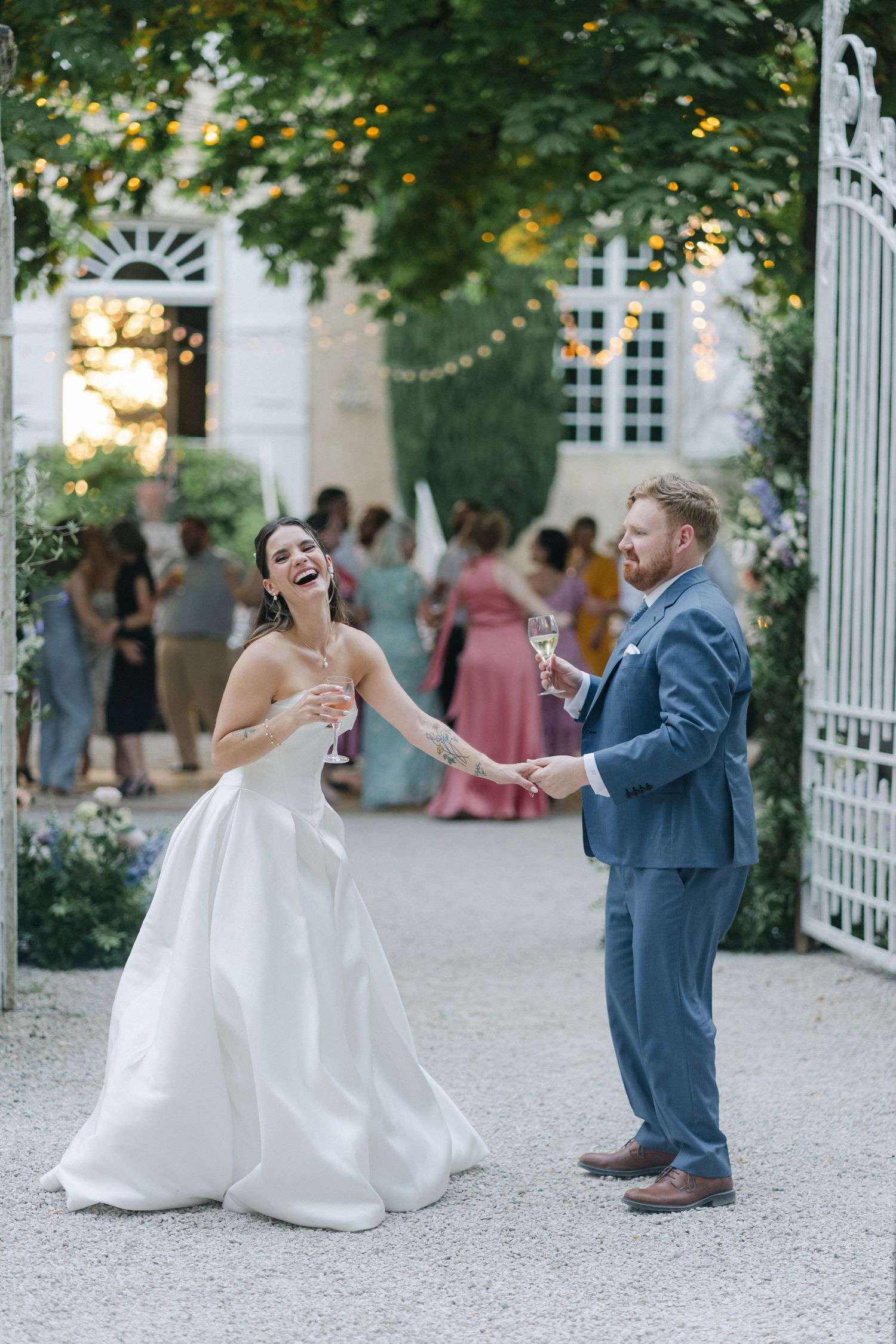 Joyful bride in white gown and groom in blue suit celebrate their wedding day outdoors with champagne and string lights.