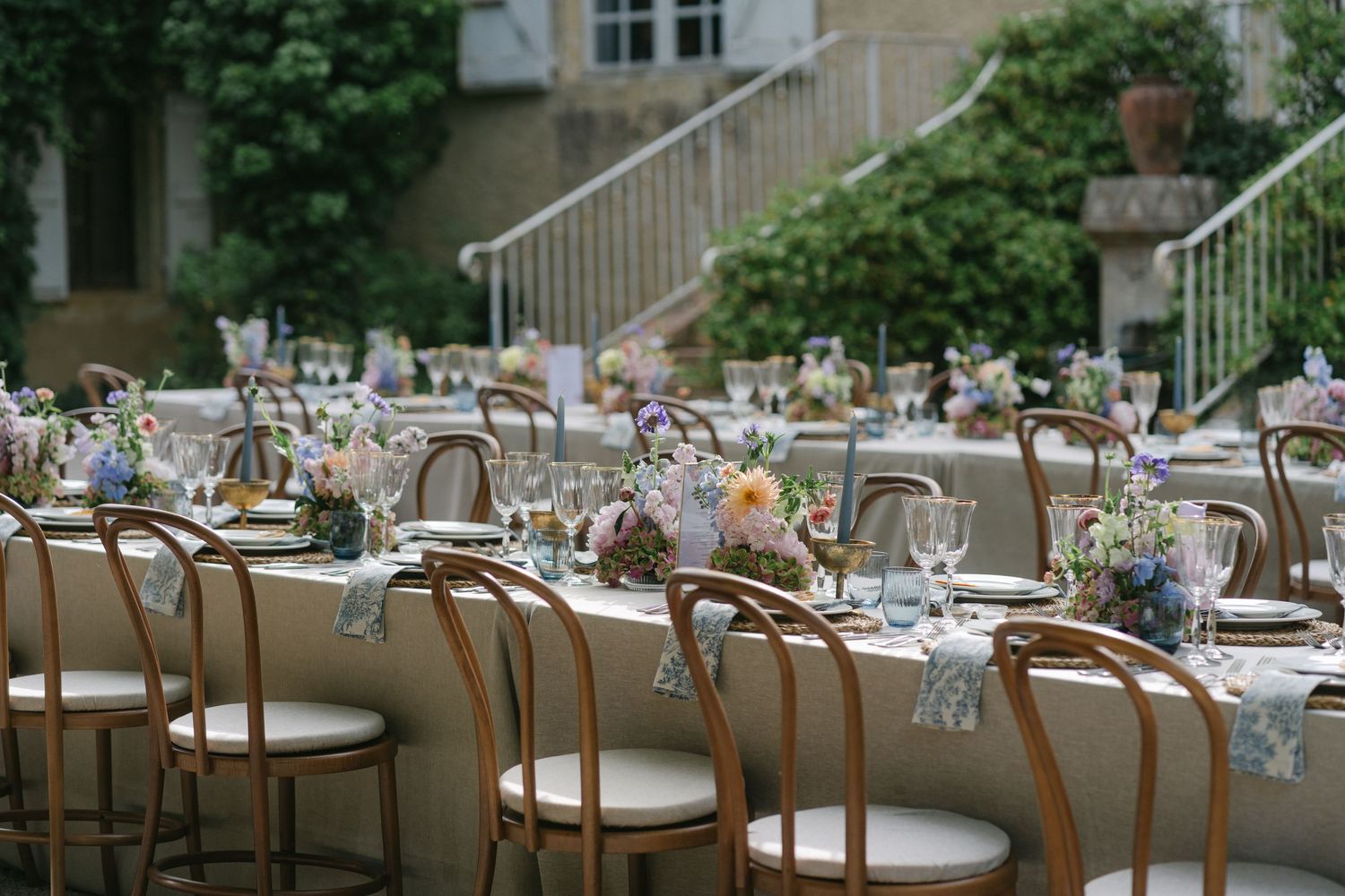 Elegant outdoor wedding reception with wooden bistro chairs, long tables, colorful floral centerpieces, and lush greenery backdrop.