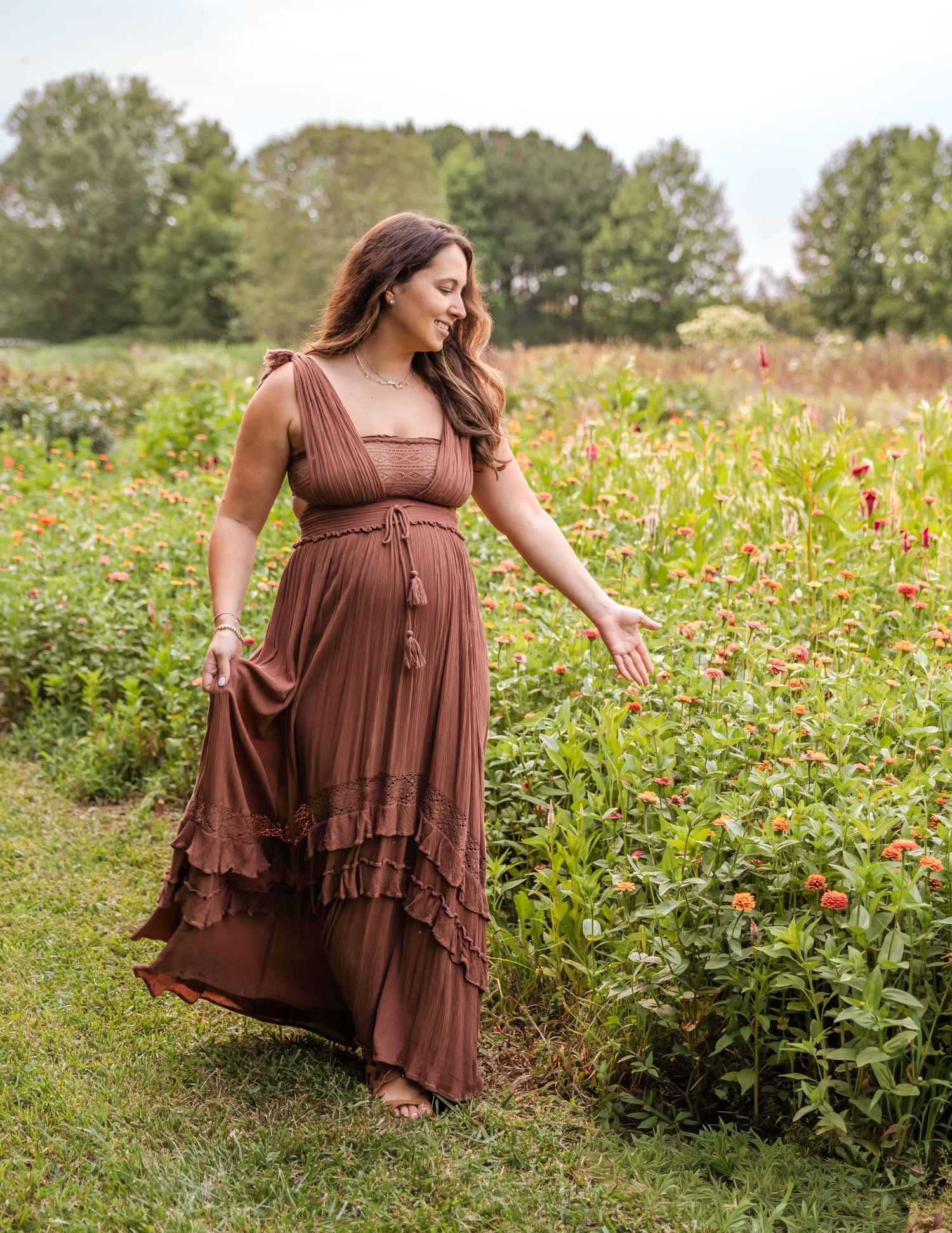 Pregnant woman in brown boho maxi dress walking through a colorful flower field outdoors.