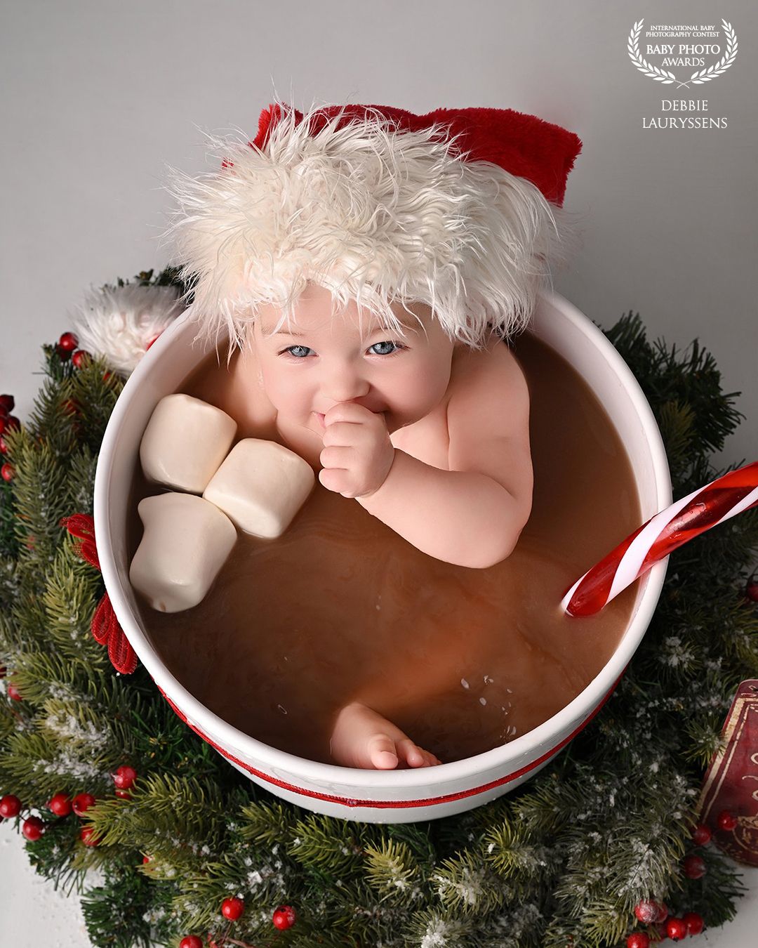 Baby in Santa hat sits in oversized hot cocoa mug with marshmallows and candy cane, surrounded by Christmas greenery.