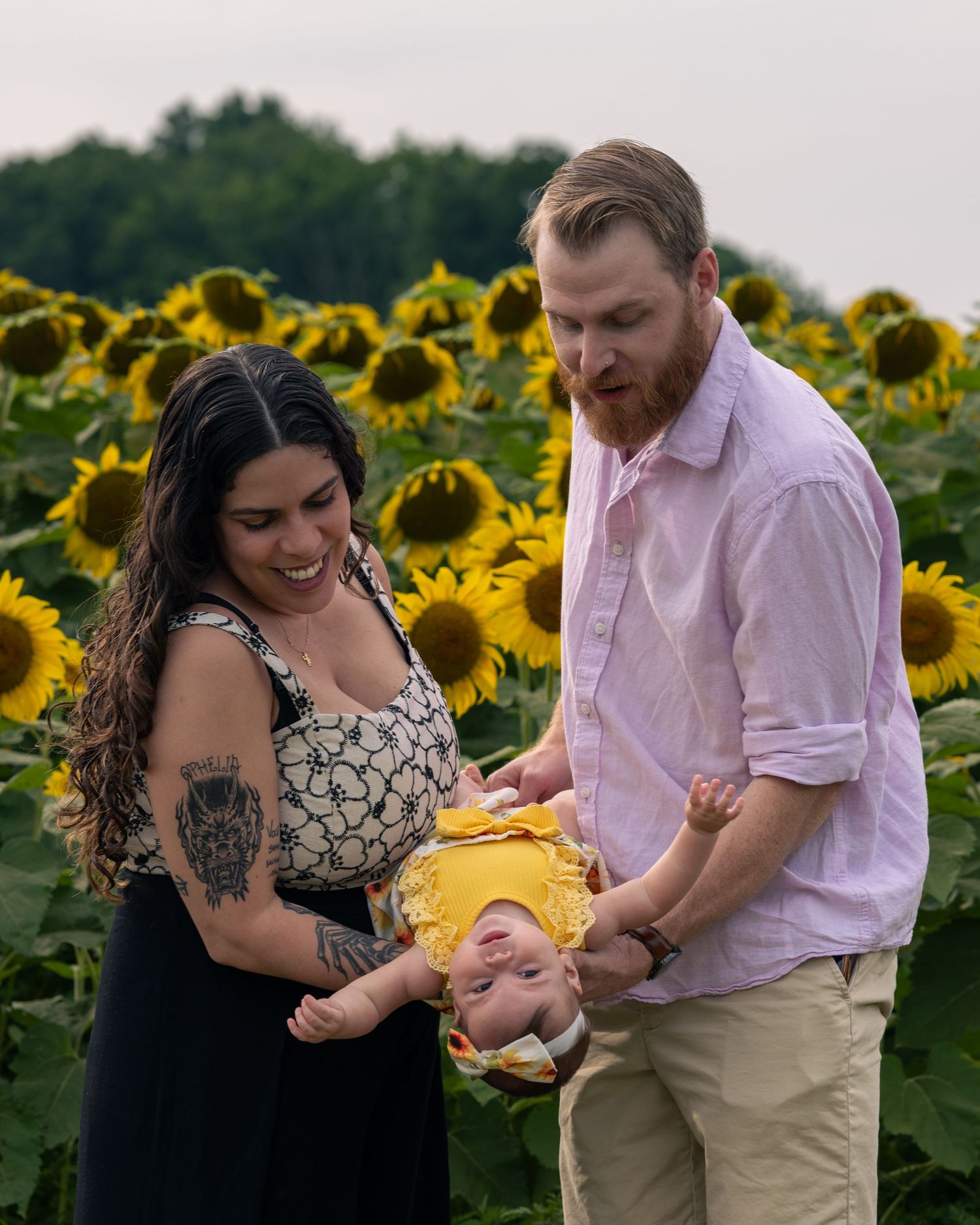 A couple holds their baby in a sunflower field, smiling joyfully during a family photo session outdoors.