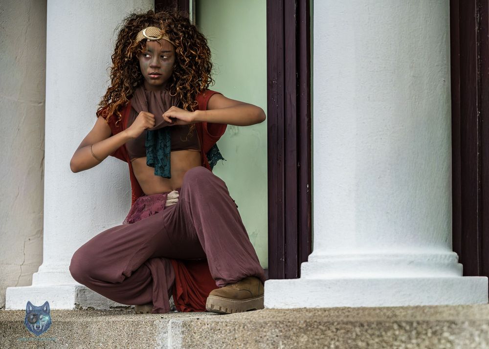 Woman in flowing burgundy pants and crop top crouching gracefully between white columns in a stylized pose.