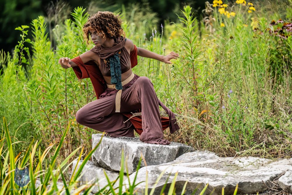 Woman in bohemian outfit balancing on a rock surrounded by lush green wildflowers in a nature setting.
