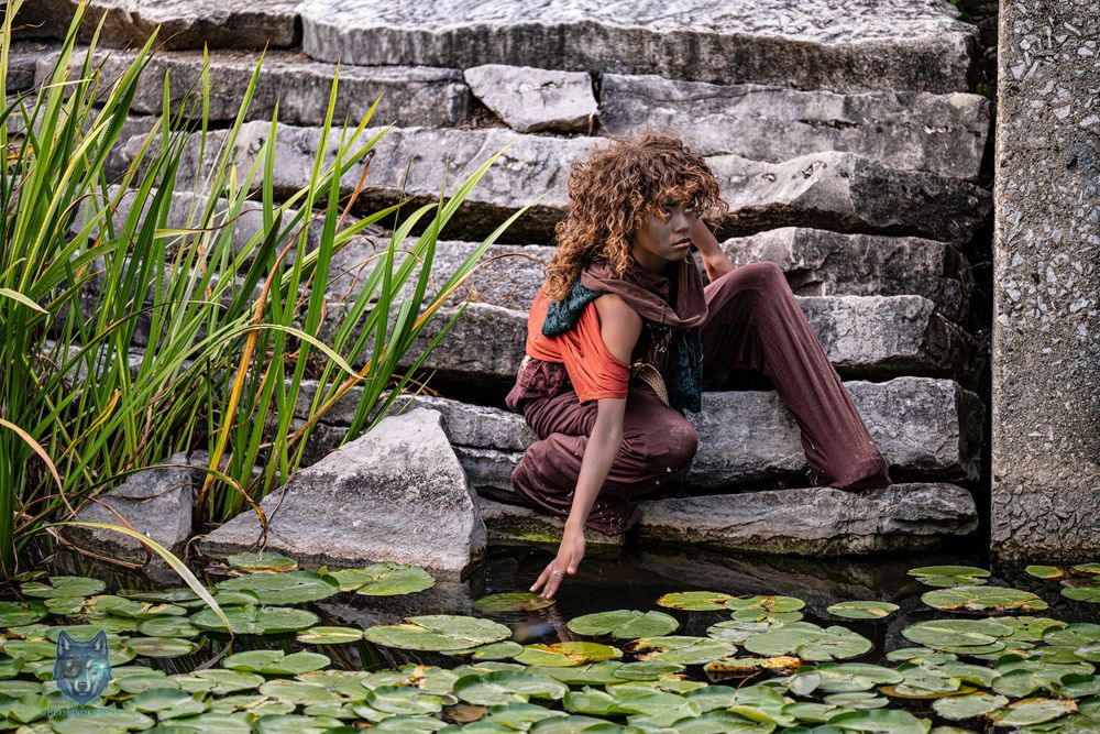 Woman in rustic costume sitting by a lily pad pond on stone steps in a fantasy-inspired photoshoot.