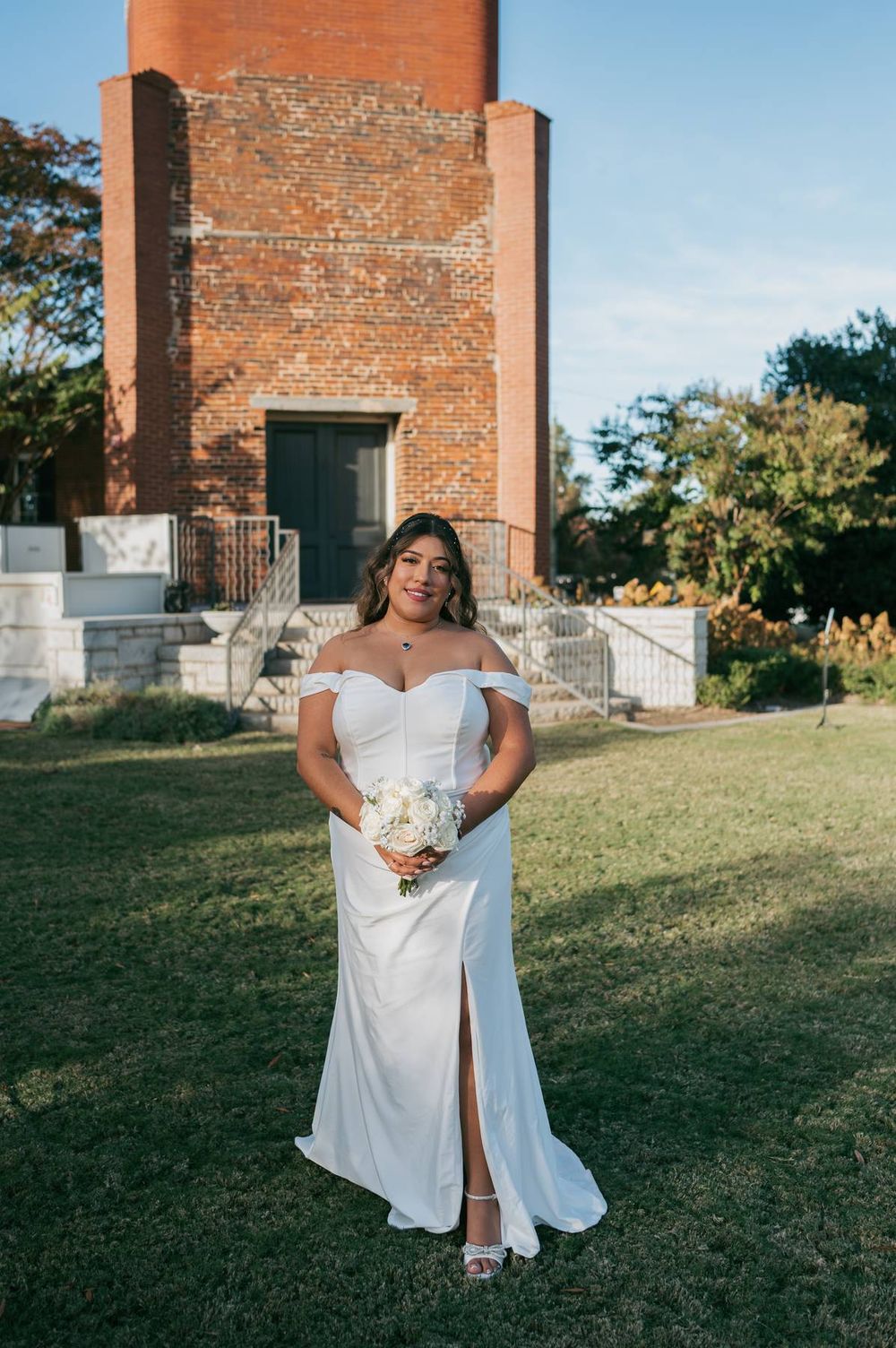 Bride in off-shoulder white gown with slit holds bouquet before historic brick tower on sunny day, wedding photographer NC