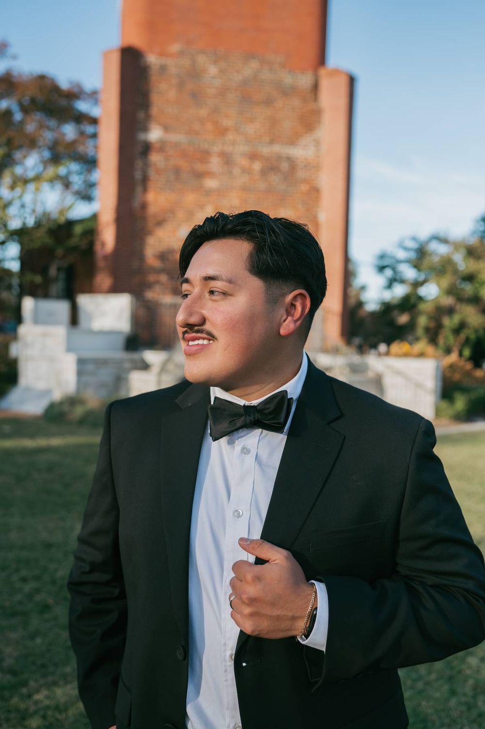 Groom in black tuxedo with bow tie stands confidently before brick tower on wedding day, wedding photographer Lexington NC