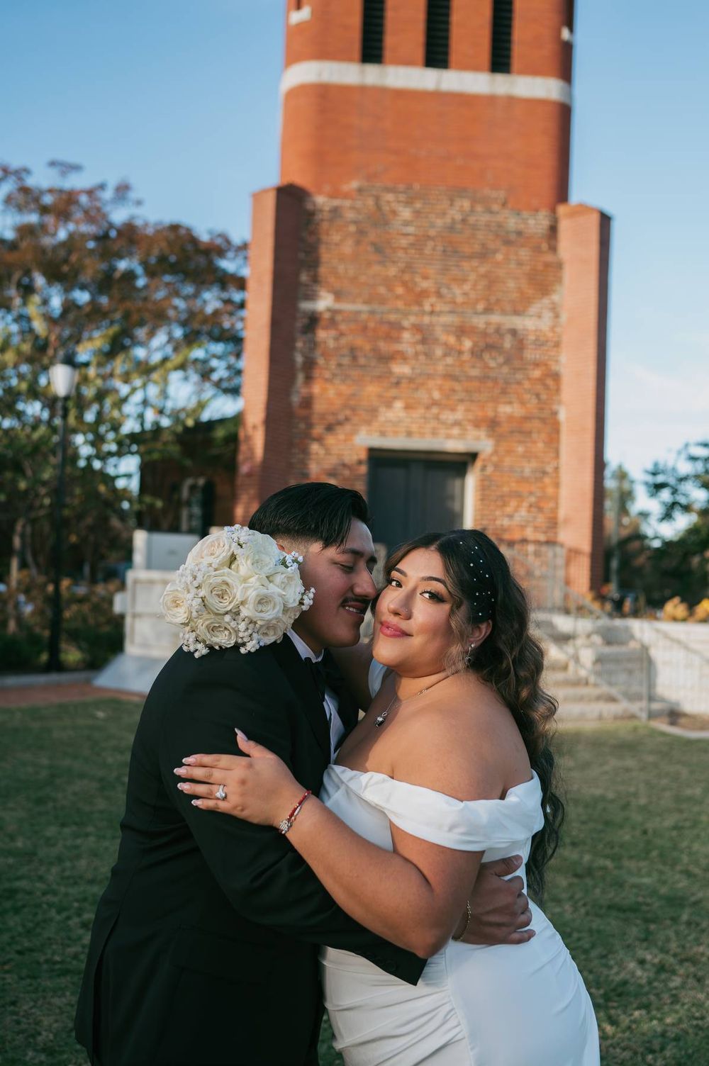 Groom embraces bride in white off-shoulder gown as she smiles at camera near brick tower, wedding photographer High Point NC