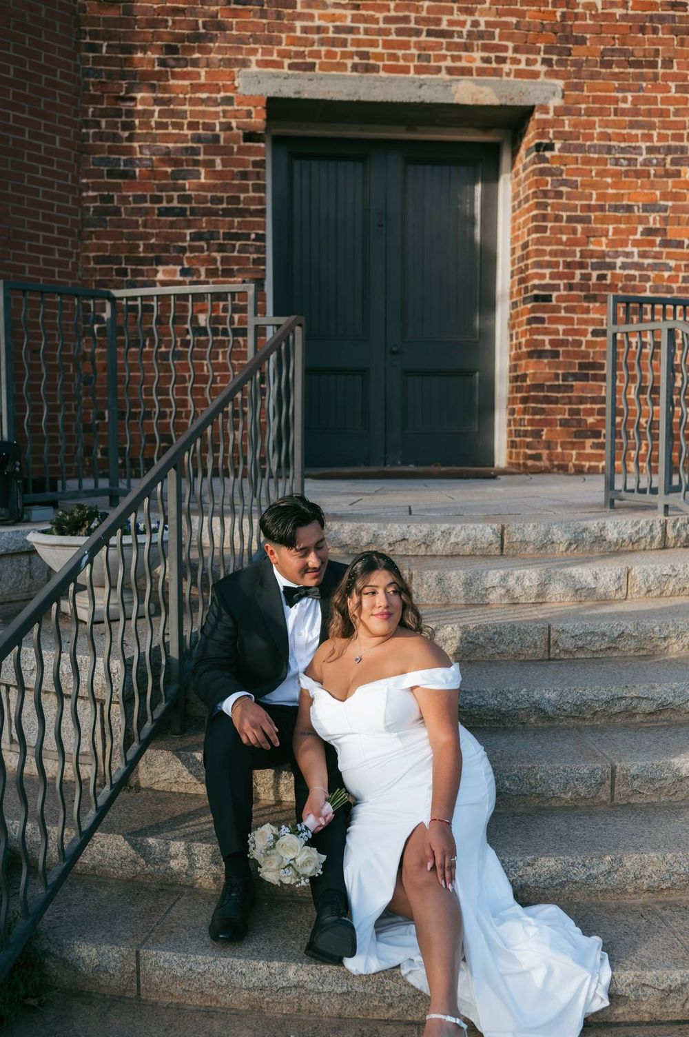 Newlyweds sit together on stone steps of brick building, bride holding white bouquet, wedding photographer Greensboro NC