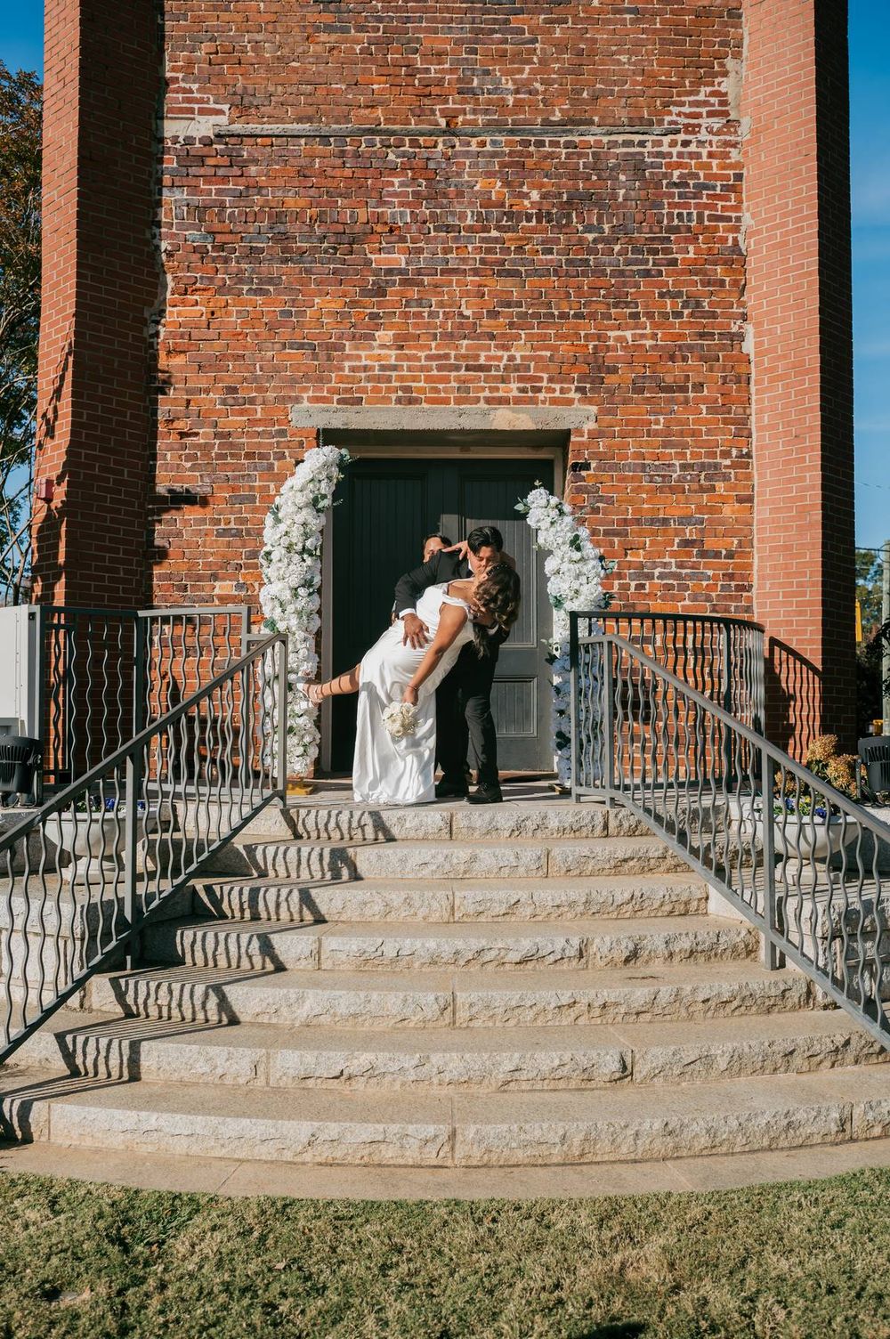 Groom dips bride dramatically at entrance of brick building adorned with white floral arrangements, wedding photographer Asheboro NC