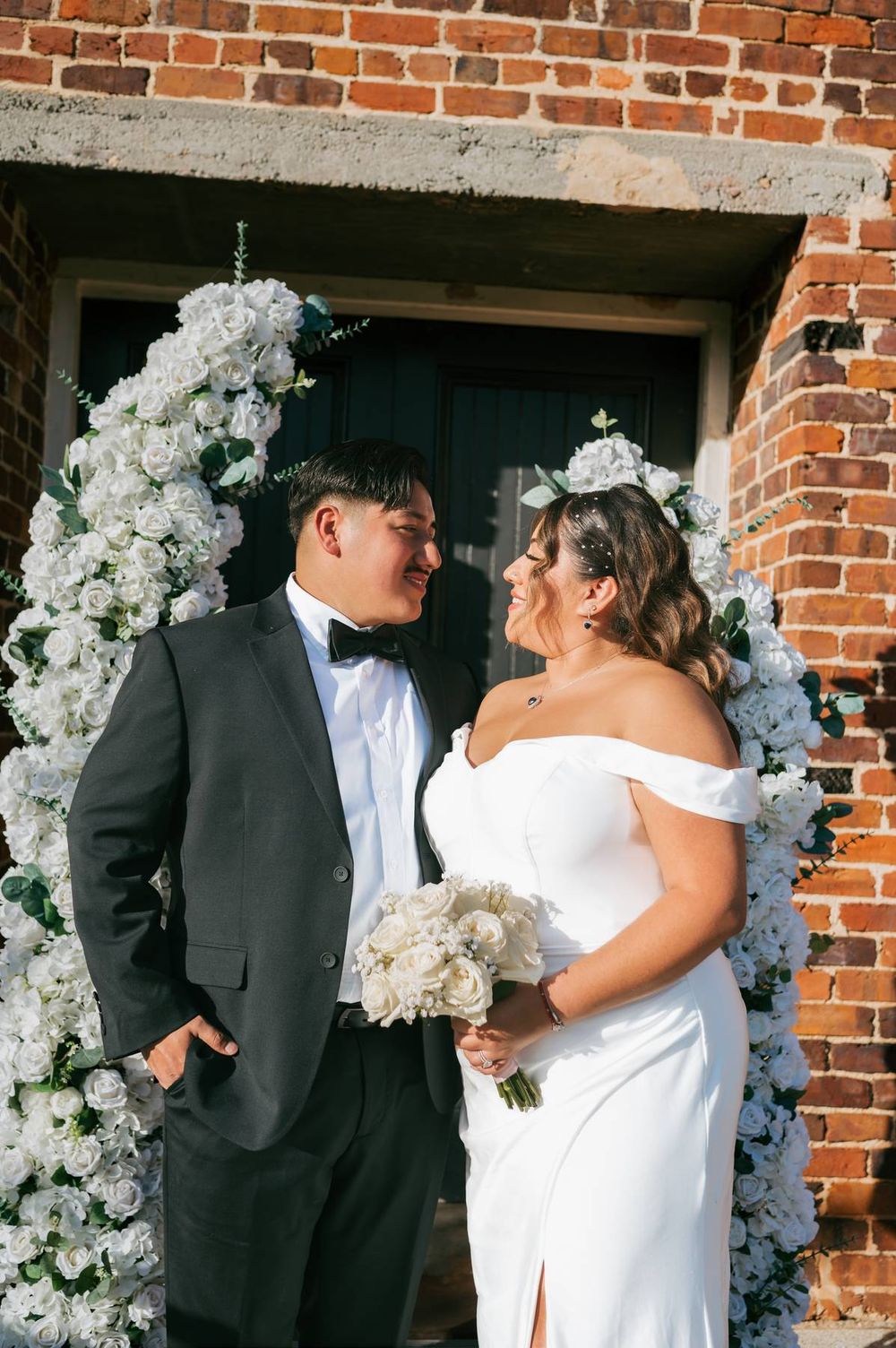 Bride and groom smile at each other under white floral arch against brick wall outdoor wedding ceremony, wedding photographer Greensboro NC