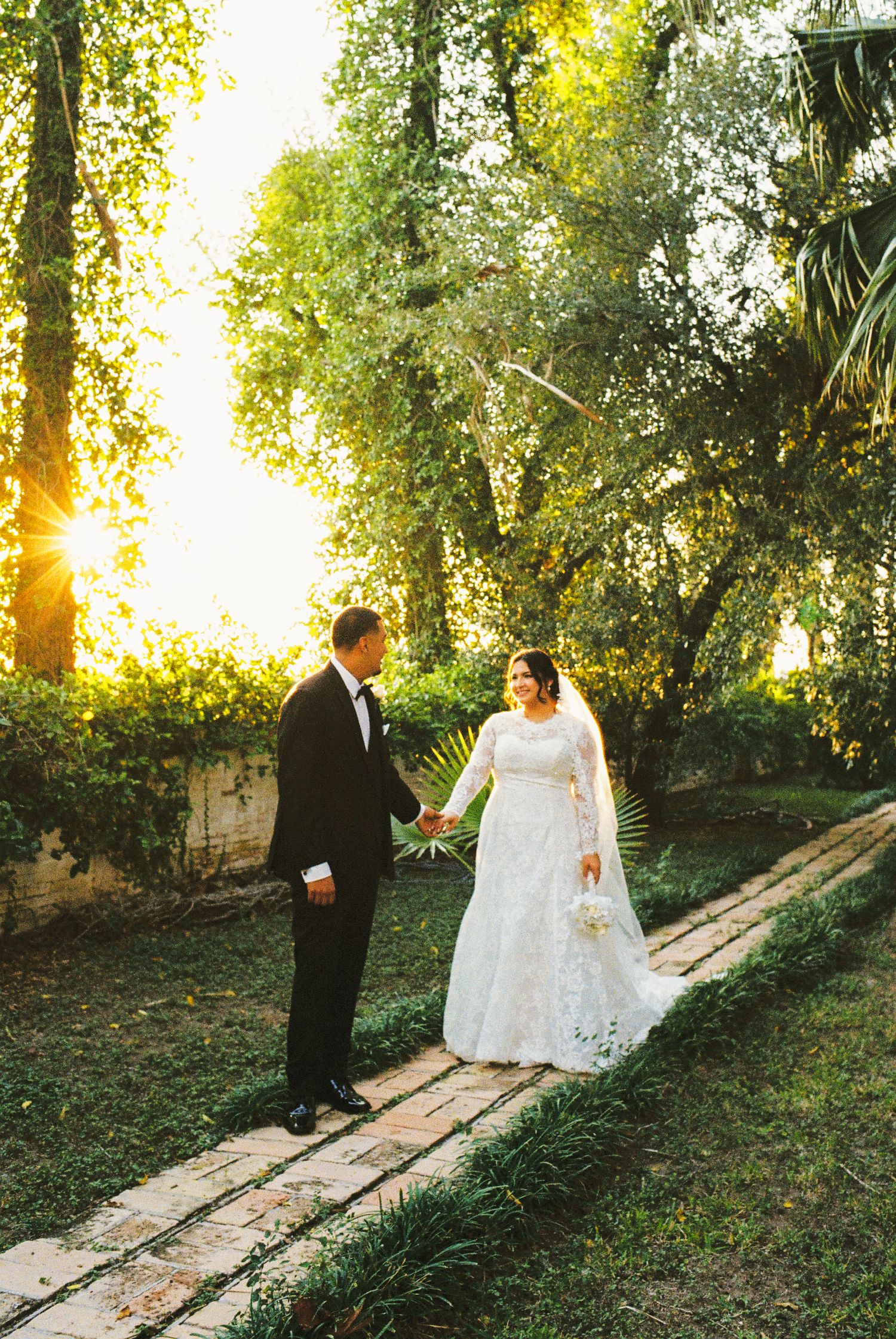 Bride and groom hold hands in a sunlit garden path, bride in lace gown, groom in black tuxedo.