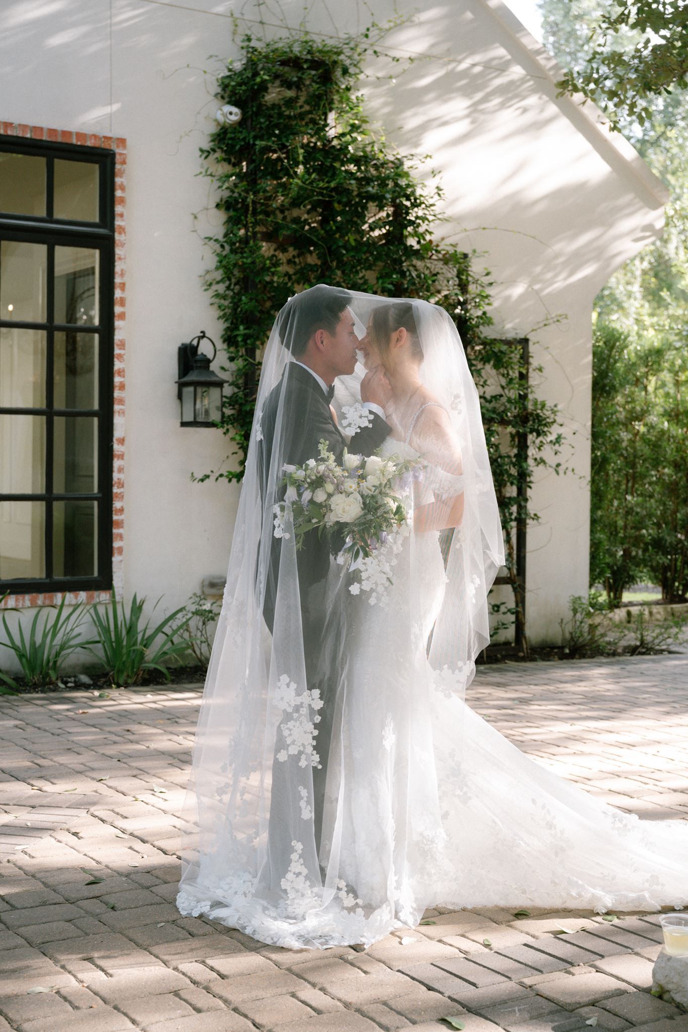 Bride and groom share a romantic moment under a flowing lace veil outdoors at an elegant white venue.