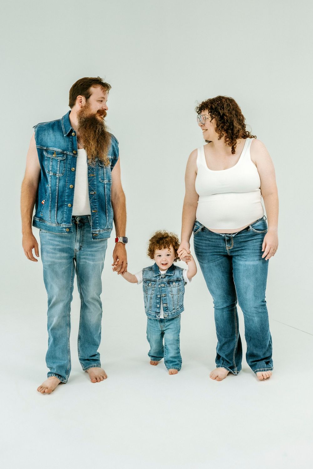 A bearded man and curly-haired pregnant woman in matching denim outfits hold hands with their toddler on a white background.