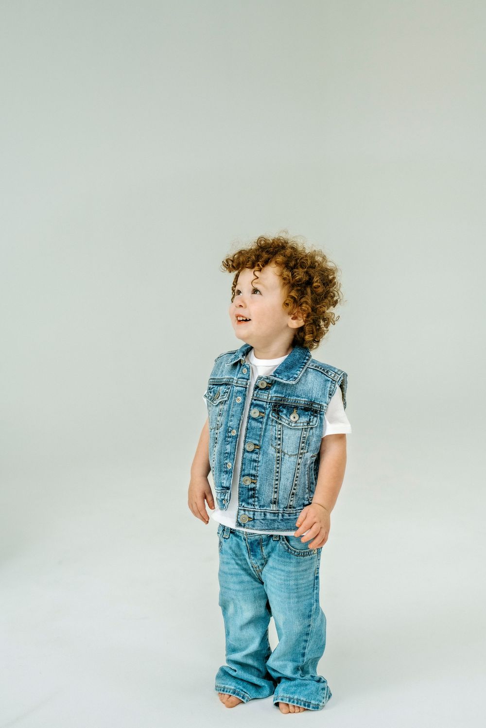 Curly-haired toddler wearing a denim vest and jeans smiling and looking up against a light gray background.