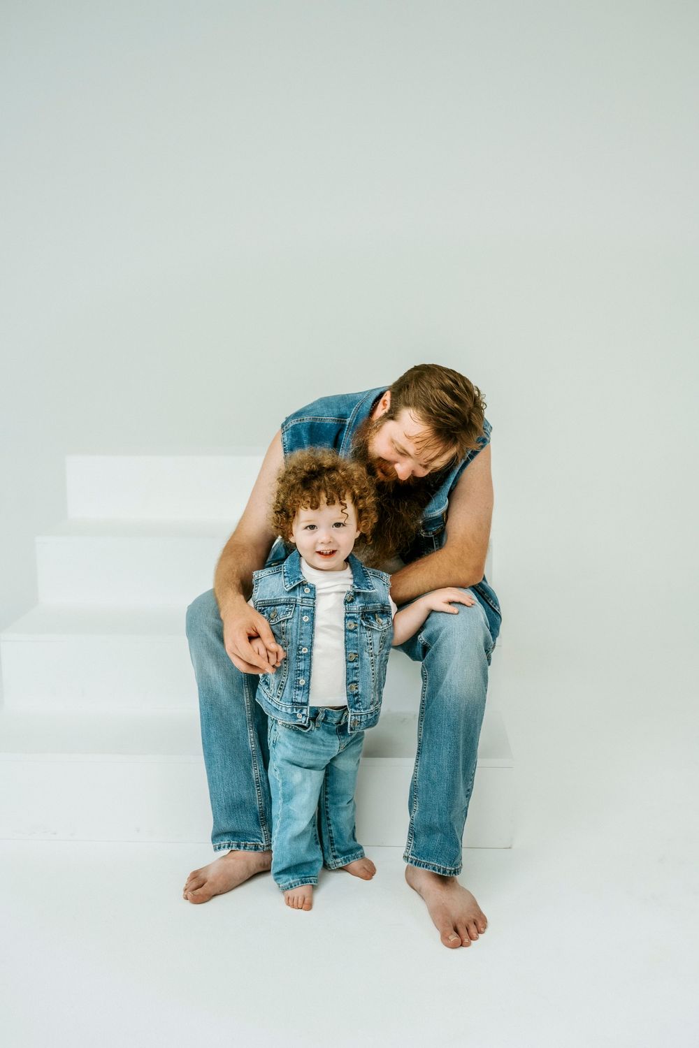 Father and curly-haired toddler in matching denim outfits sitting on white steps, smiling together.