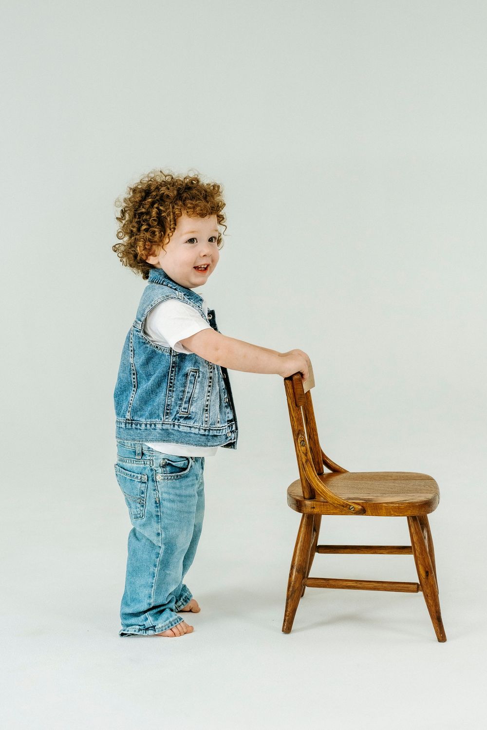 Curly-haired toddler in denim vest and jeans standing beside a wooden chair on a white background.