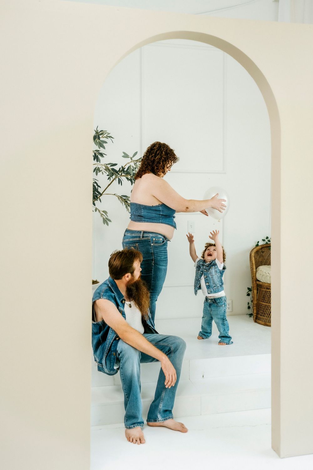 Family in denim outfits playing together in a bright white room with an arched doorway and indoor plants.