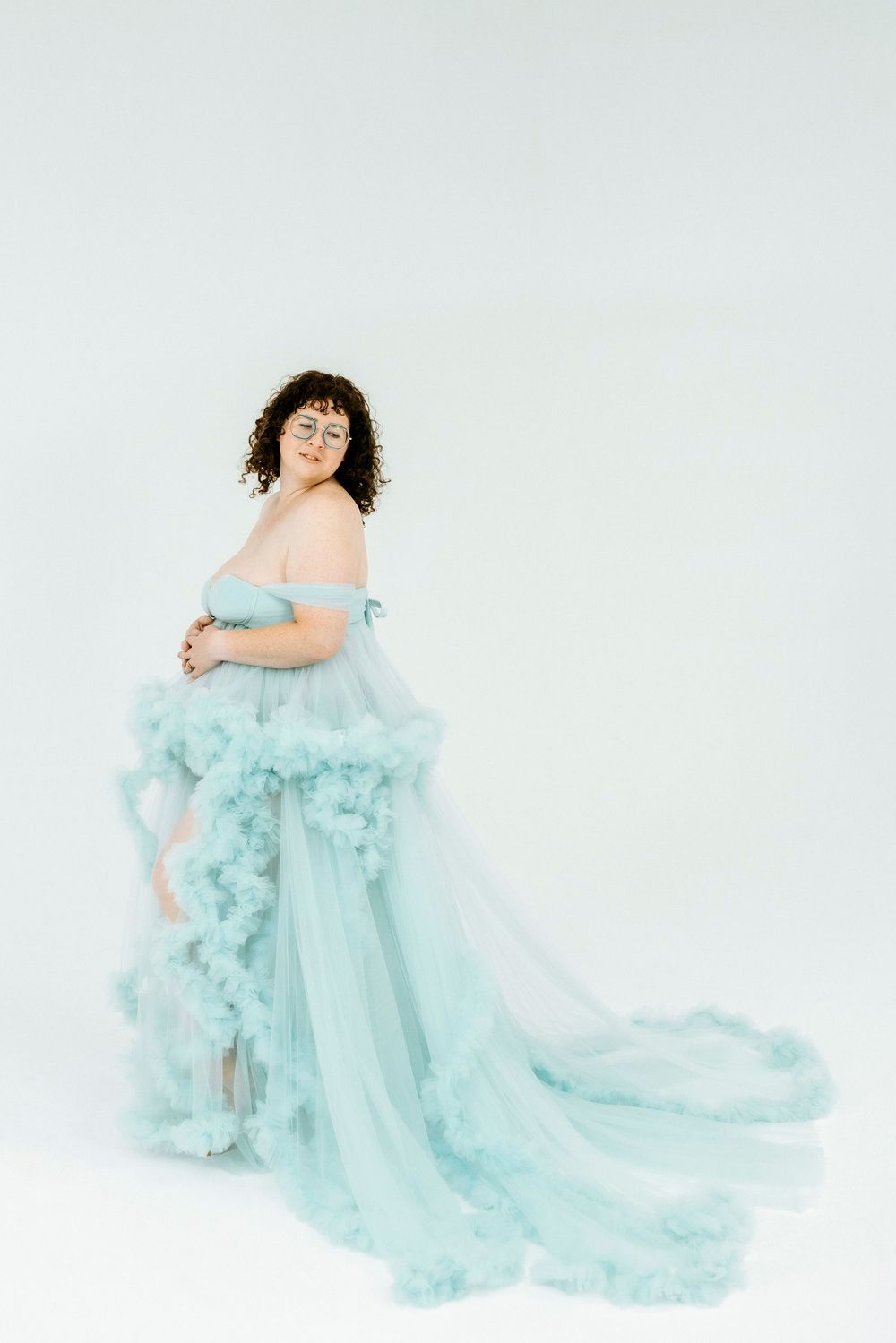 Pregnant woman in elegant off-shoulder teal tulle maternity gown posing gracefully against white background.