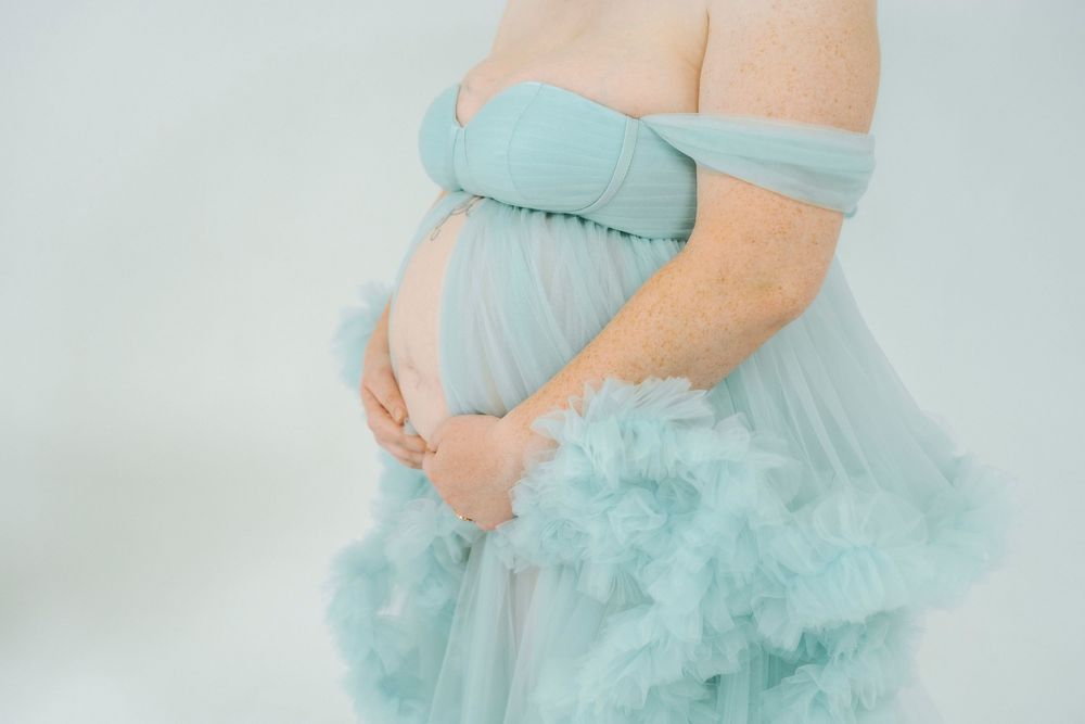 Pregnant woman in elegant off-shoulder light blue tulle maternity gown cradling baby bump against white background.