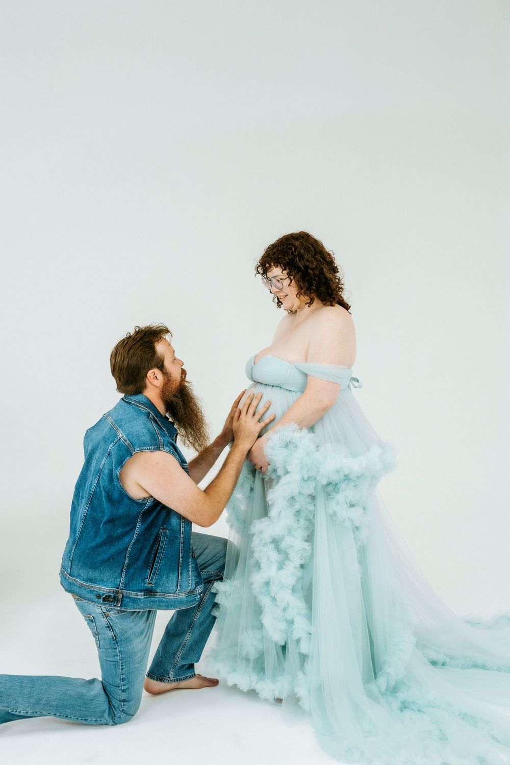 A bearded man kneels before a pregnant woman in a flowing teal tulle gown during a maternity photo session.
