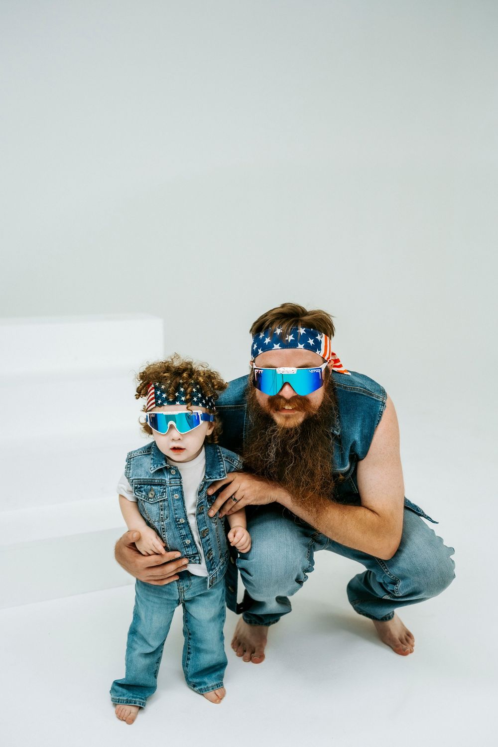Father and toddler wearing matching American flag headbands and colorful sunglasses in denim vests pose together on white background.