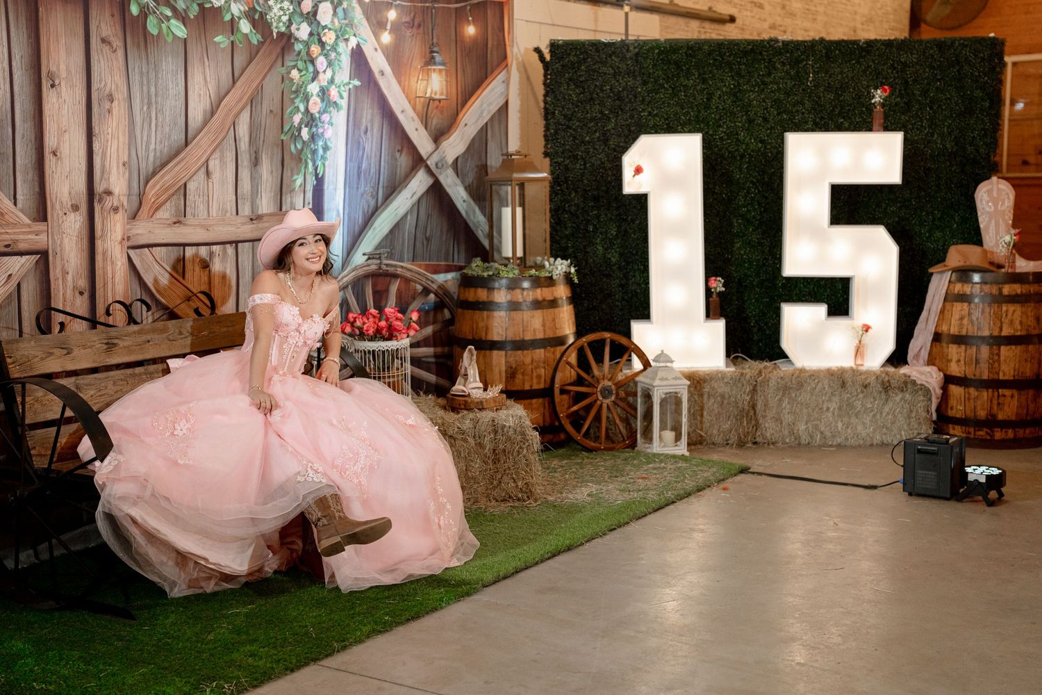 A young woman in a pink ball gown poses at a rustic quinceañera setup with a large number 15 display and barn decor.