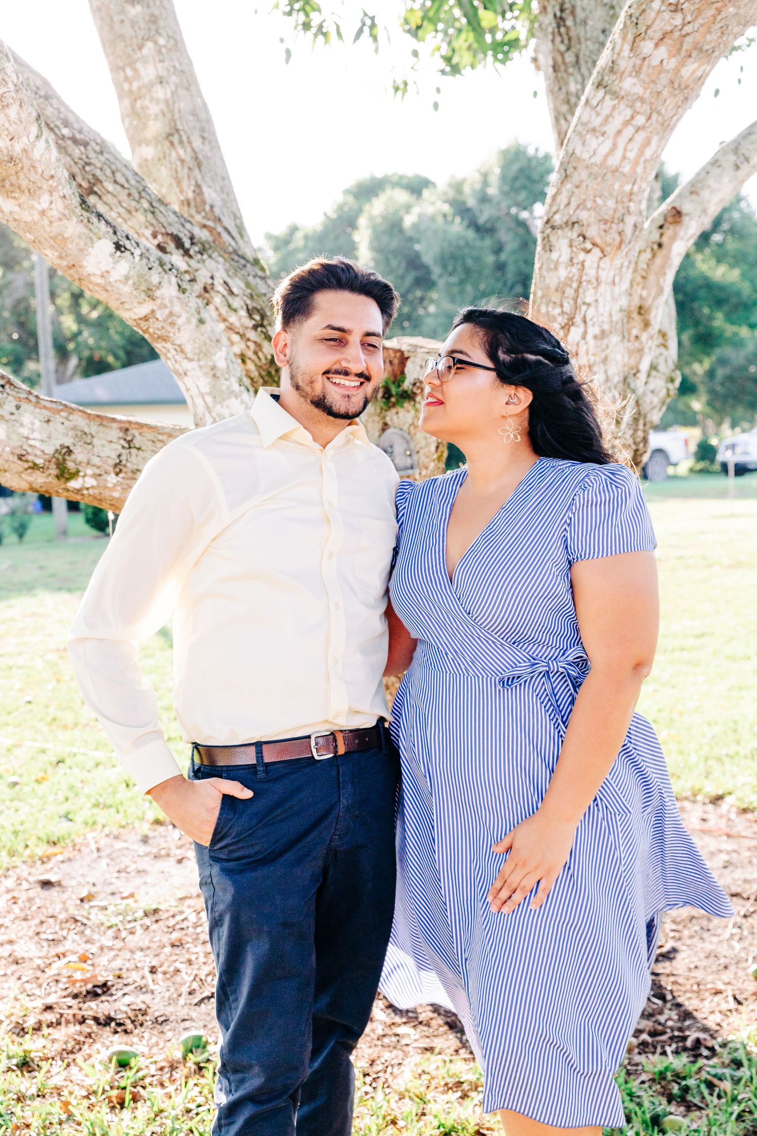 Happy couple smiling at each other outdoors under a tree, man in white shirt and woman in blue striped dress.