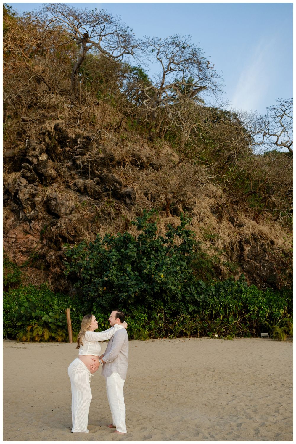 One & Only Mandarina family photographer showcasing maternity photos with a couple embracing on a beach surrounded by cliffs and greenery.