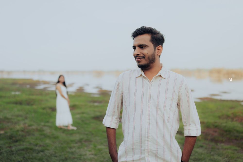 A smiling man in a striped shirt stands outdoors near a lake, with a woman in a white dress in the background.