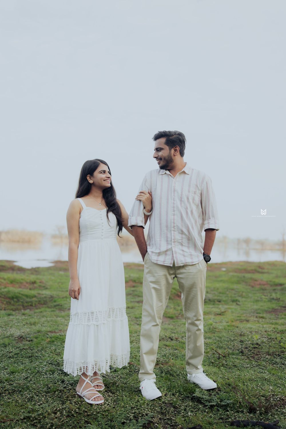 A couple in white outfits smiling at each other outdoors near a lake on a cloudy day.
