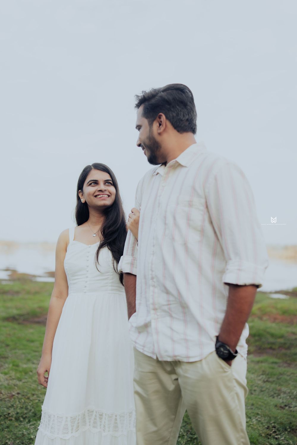 A smiling couple in white outfits standing outdoors near water, sharing a tender moment on a cloudy day.