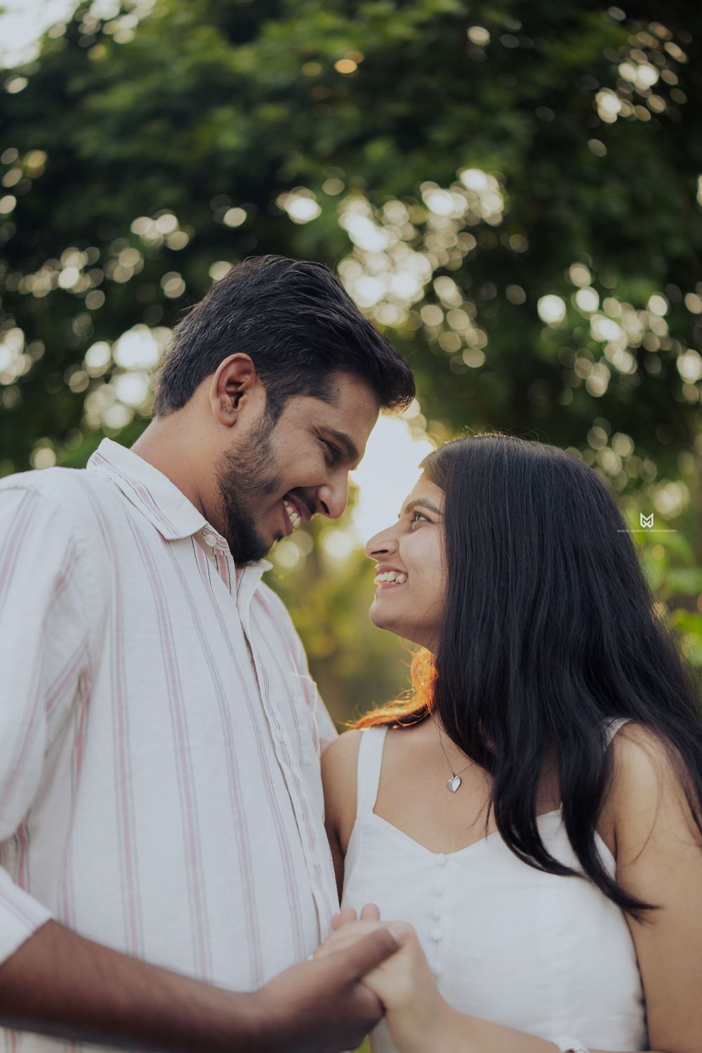 A smiling couple gazes lovingly at each other outdoors, with soft bokeh green trees in the background during golden hour.