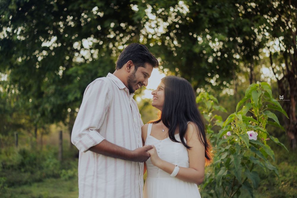 A couple shares a romantic moment outdoors, surrounded by lush green trees and blooming flowers.
