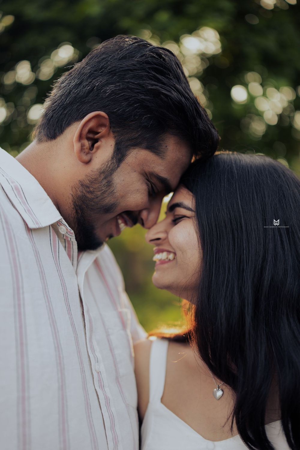 A couple sharing a romantic forehead-to-forehead moment outdoors, smiling warmly with bokeh green trees in the background.