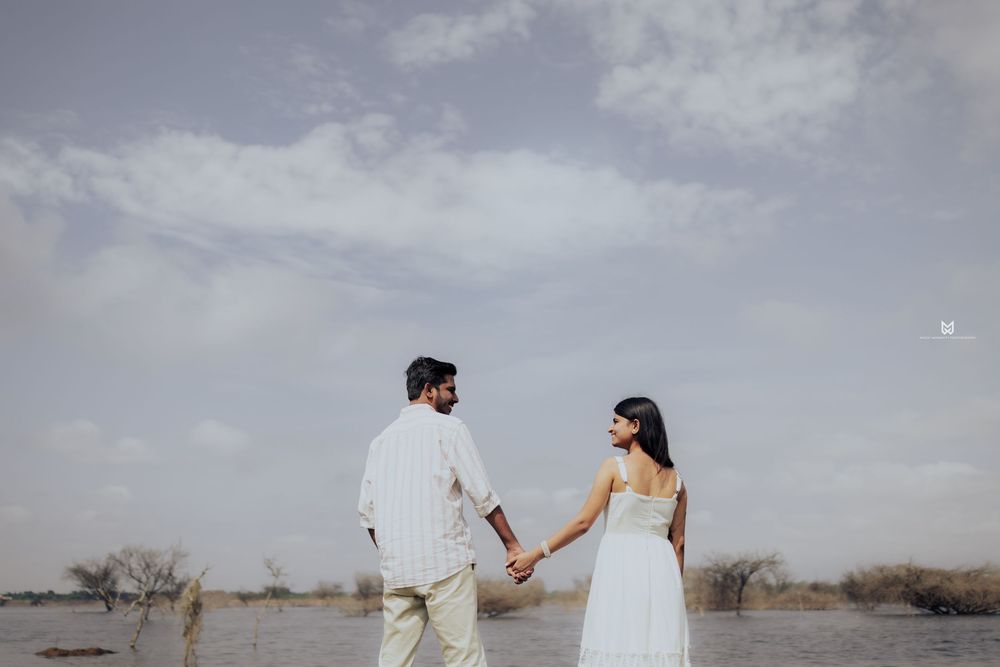 Couple holding hands facing a serene lake under a cloudy sky, woman in white dress, man in casual white outfit.