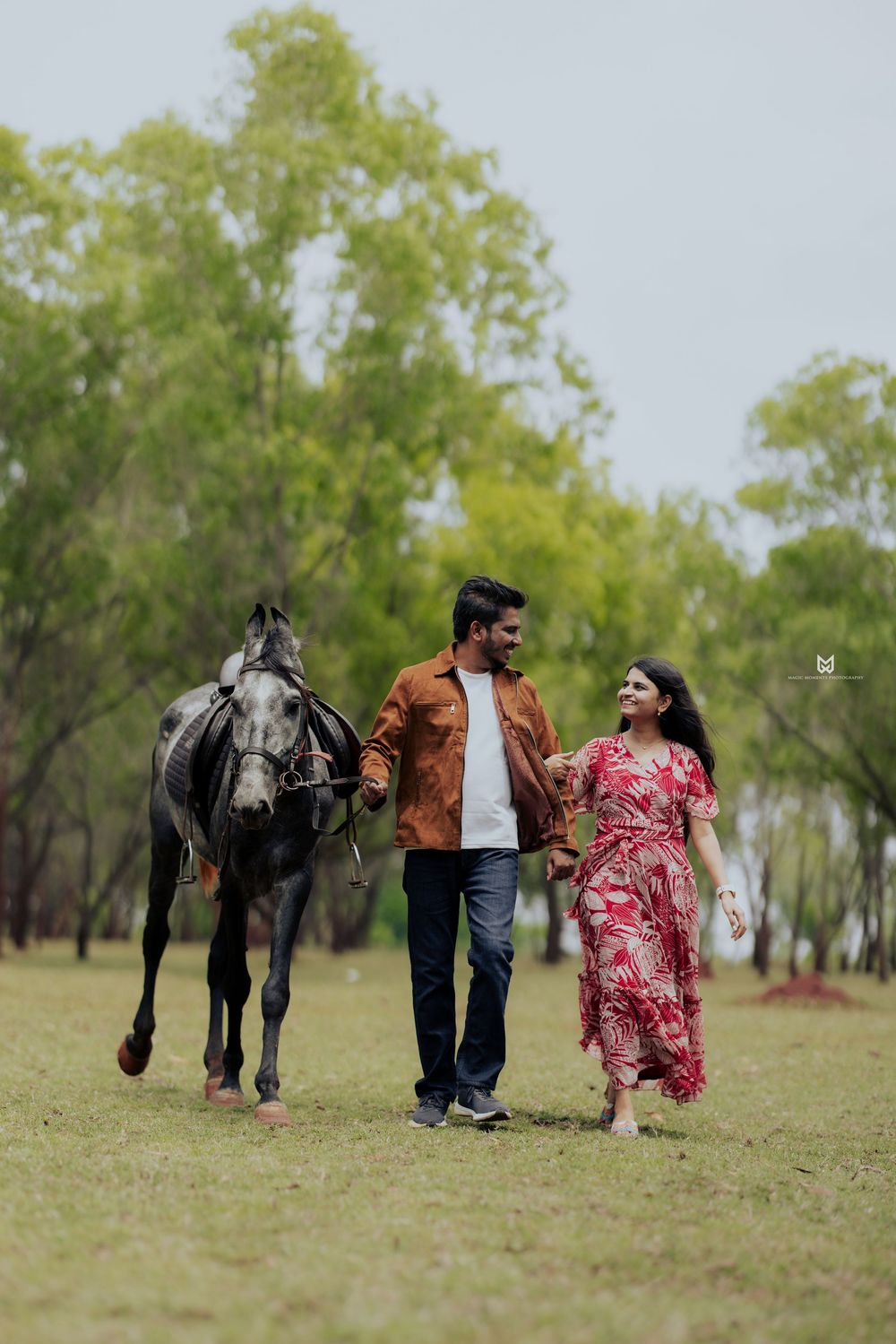Couple walking with a dark horse in a lush green park, woman in red floral dress, man in brown jacket.