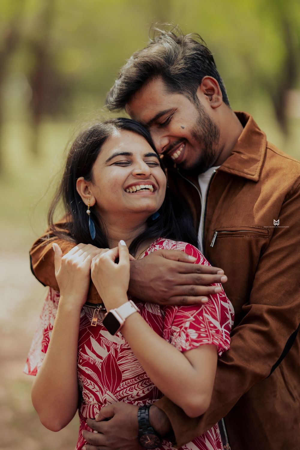A joyful Indian couple embracing outdoors, woman in floral dress laughing, man in brown jacket smiling, green trees background.