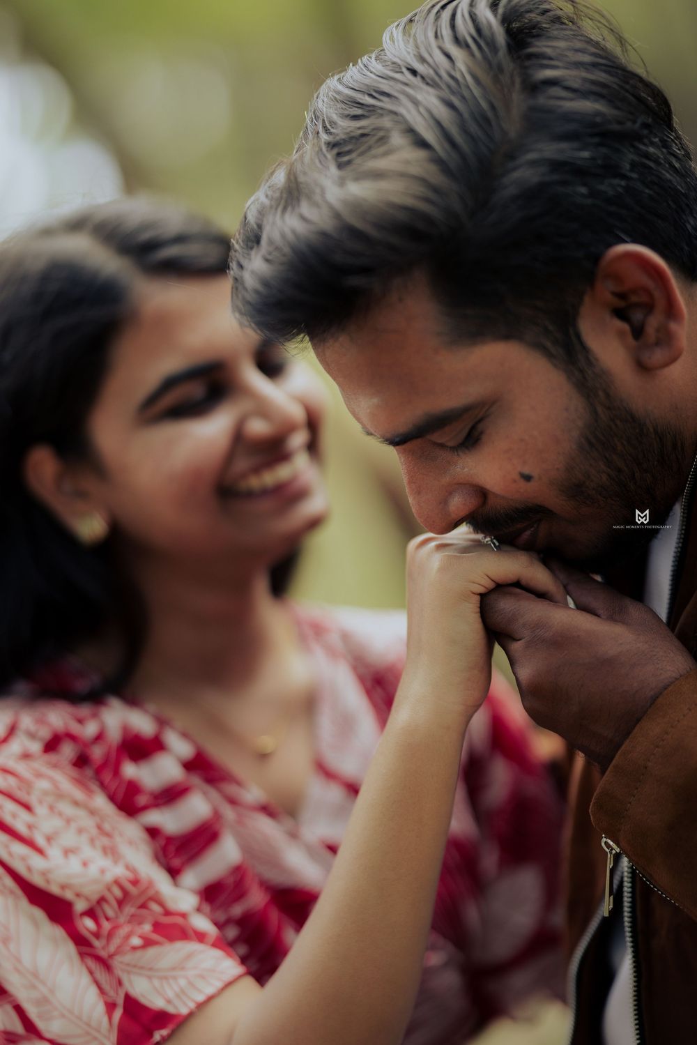 A man tenderly kisses a woman's hand while she smiles warmly in a romantic outdoor couple portrait.
